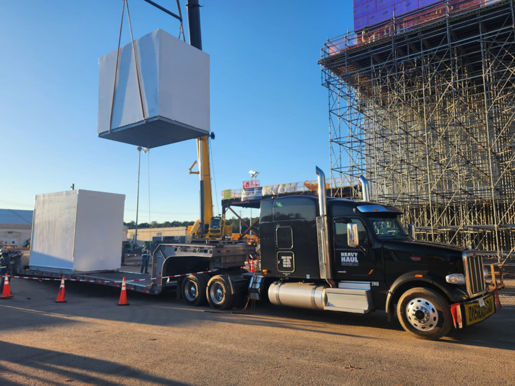 Crane lifting a modular equipment enclosure onto a trailer for heavy haul transport and delivery.