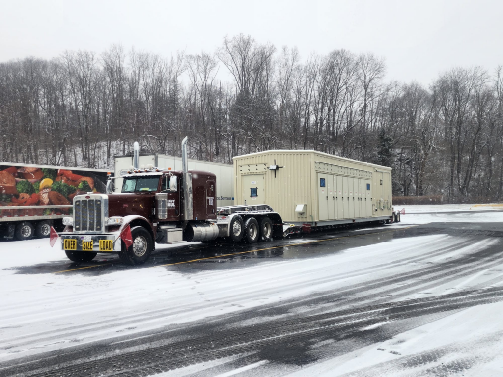 Oversize load heavy haul truck hauling a large beige electrical equipment shelter.