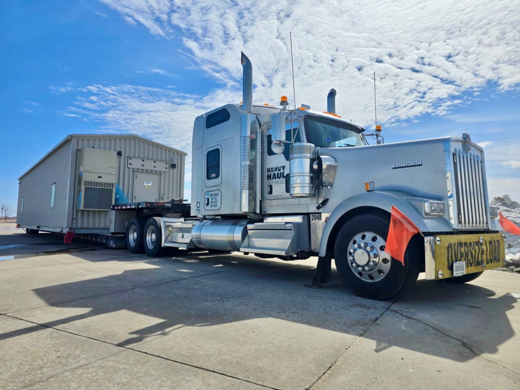 Silver Kenworth heavy haul truck with OVERSIZE LOAD banner hauling a prefabricated unit.